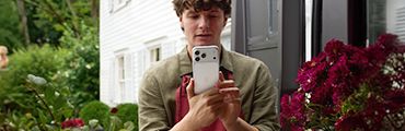 Person hält Smartphone vor Gesicht, weißes Gebäude, rote Blumen rechts